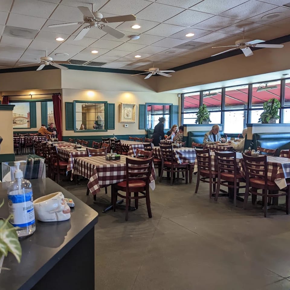 Bright restaurant dining room with checkered tablecloths, blue booths, ceiling fans, large windows, and several diners.