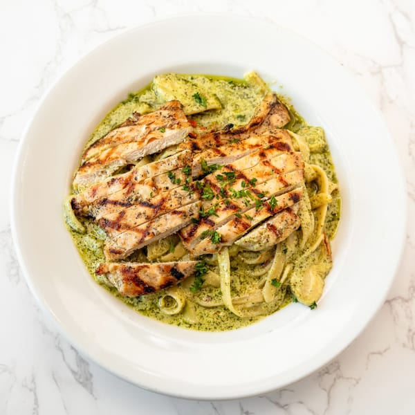 Grilled chicken slices over fettuccine pasta with green pesto sauce in a white bowl.
