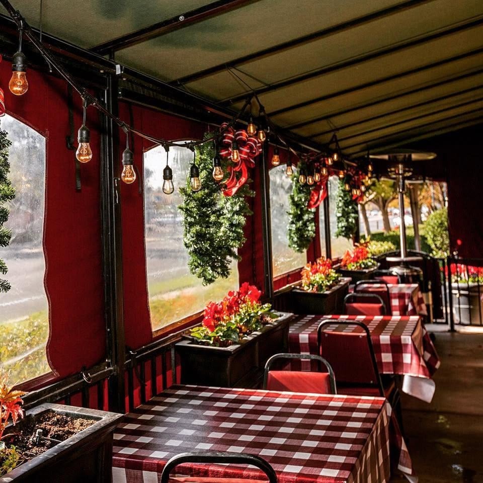 Festive restaurant patio with red-and-white checkered tables, hanging Edison bulbs, greenery, and holiday decorations.