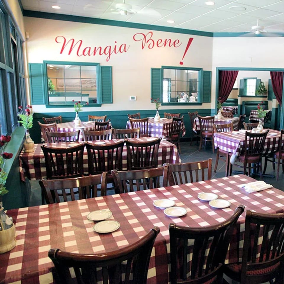 Manggia Bene restaurant dining room with checkered tablecloths, wooden chairs, floral centerpieces, and Italian-style decor.