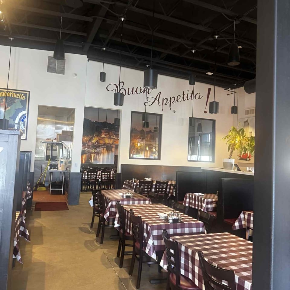 Italian restaurant dining room with red-and-white checkered tablecloths, black chairs, pendant lights, and “Buon Appetito!” wall lettering.