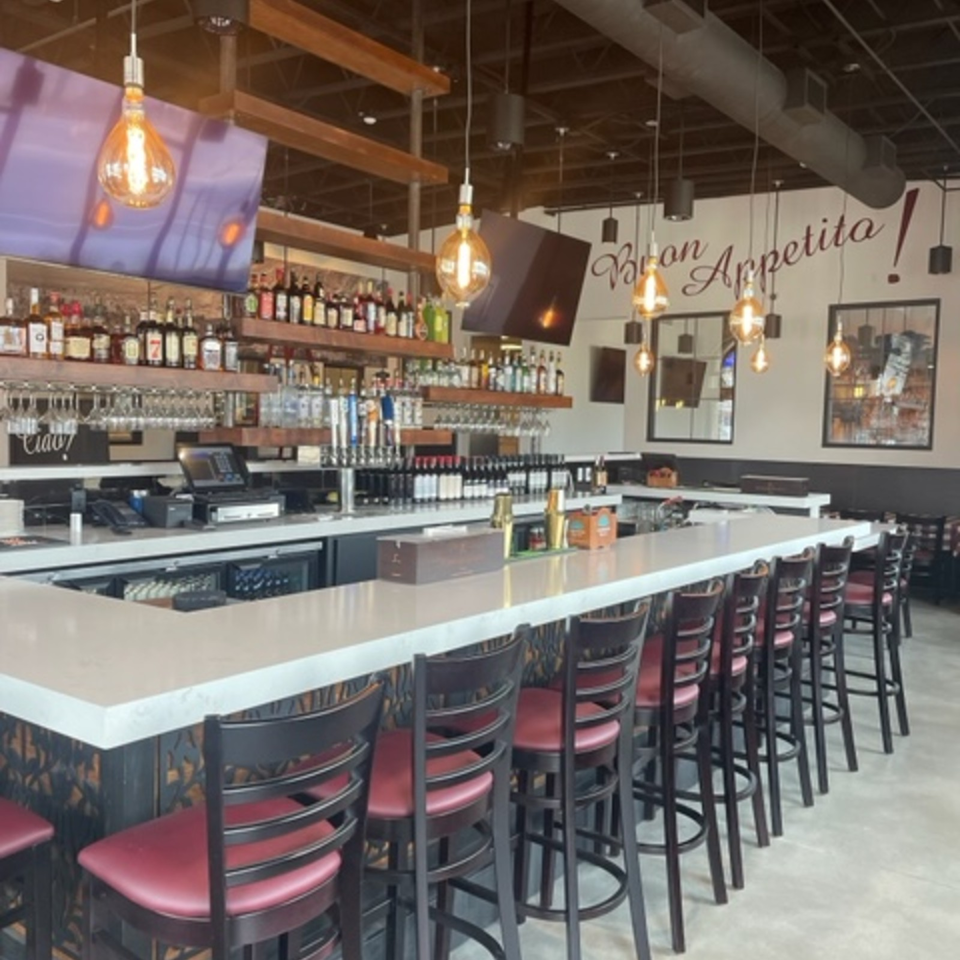 Restaurant bar with liquor shelves, pendant lighting, red leather stools, and a wall reading “Buon Appetito!”