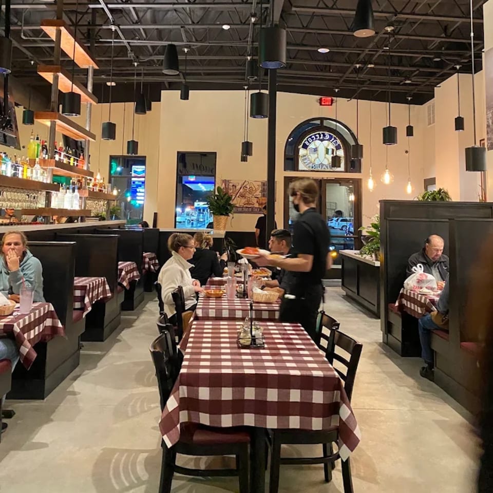 Bustling restaurant dining room with checkered tablecloths, seated diners, a server carrying food, bar, and pendant lighting.