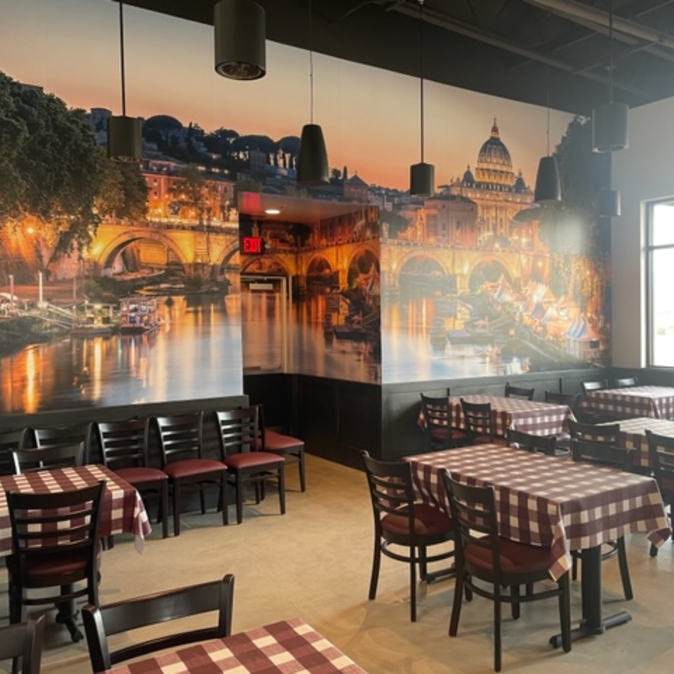 Restaurant dining room with red-and-white checkered tablecloths, pendant lights, and a sunset mural featuring St. Peter’s Basilica.