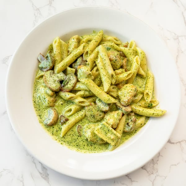 Penne pasta with chicken and mushrooms in a vibrant green pesto cream sauce, served in a white bowl.