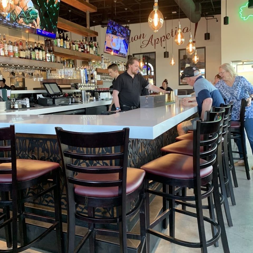 Bright restaurant bar with liquor shelves, pendant lights, high-top seating, bartender, and two patrons.